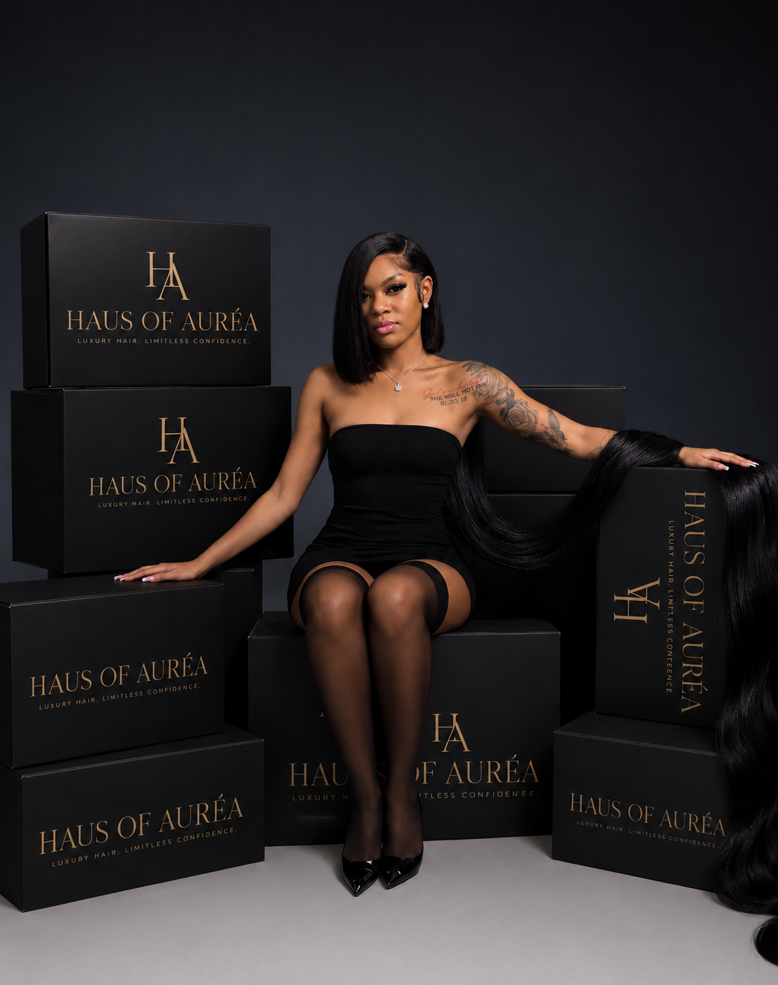 A woman in a black strapless dress and sheer black tights sits among stacked black HAUS OF AURÉA boxes, with a long black wig draped over one box in a dark studio setting.